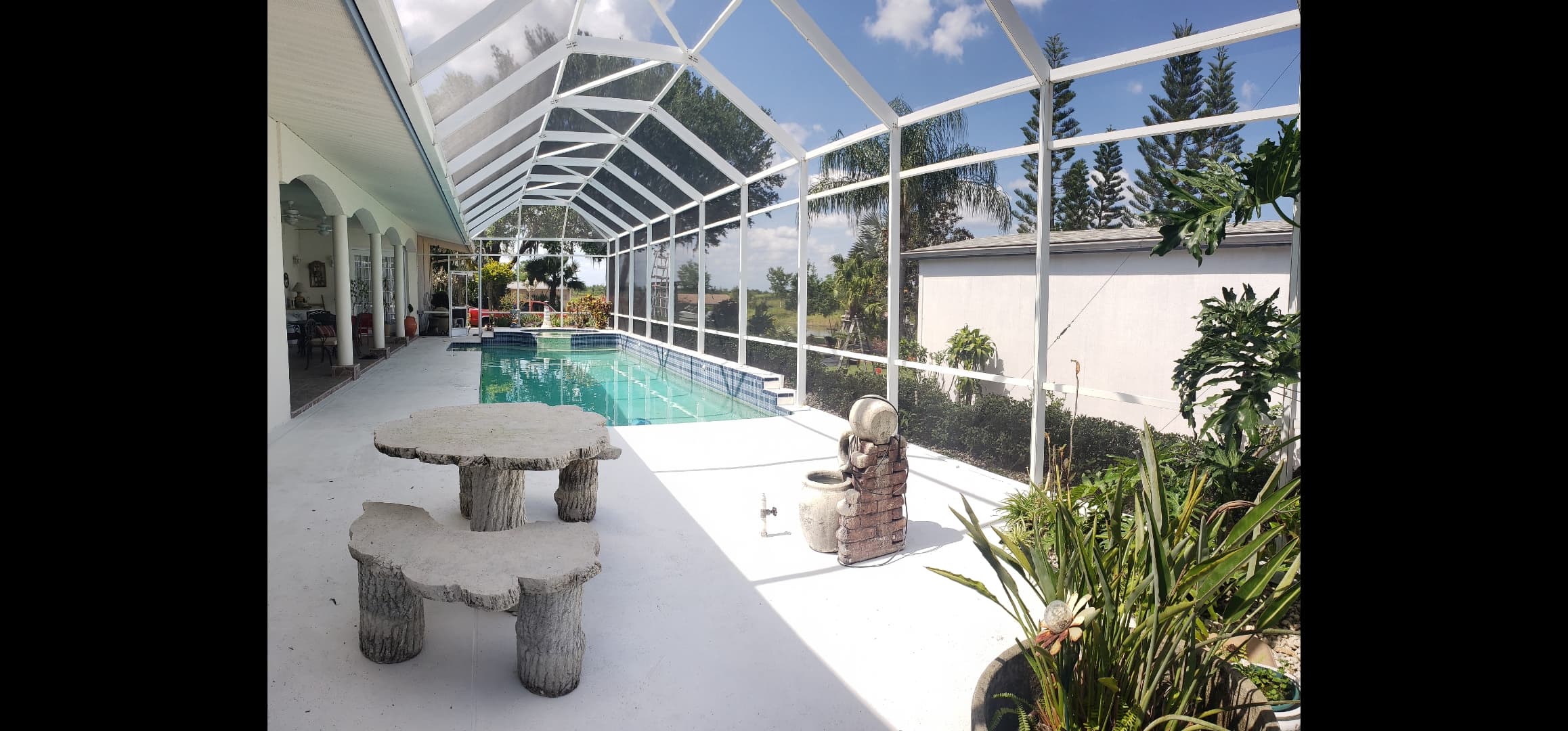 Swimming pool inside a white screen enclosure with stone furniture and lush tropical plants.